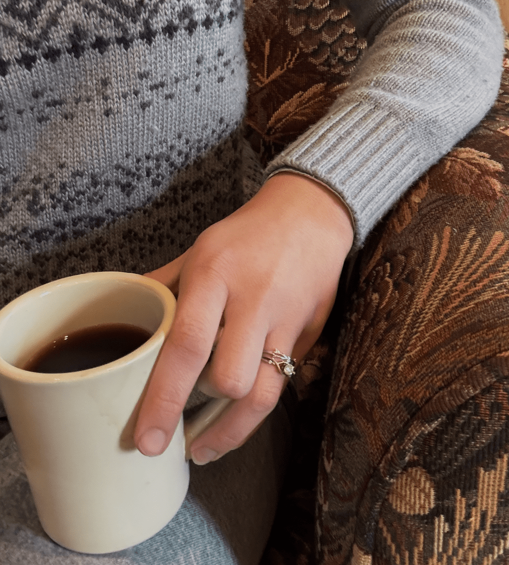 a small, diner mug of black coffee is held by  someone at Campfire Bay Resort's coffee shop. We can see the corner of her sweater and the sleeve - it's a grey-blue Norwegian pattern. She wears a set of wedding rings.